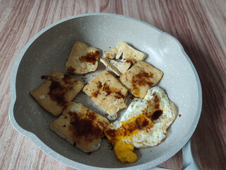 A close-up overhead shot shows a speckled gray frying pan containing several pieces of pan-fried tofu and a fried egg. The tofu is golden brown and glistening with a dark sauce. The egg yolk is still 