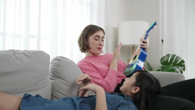 Asian cute daughter listening acoustic music while lie on her mother lap at sofa. Caucasian mother playing colorful ukulele while cute child looking at her parent. Family recreation concept. Pedagogy.