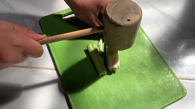 A hand uses a wooden mallet to crush an ingredient on a green cutting board.