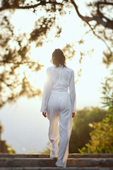 Rear view of a stylish woman in a white outfit walking toward a sunlit path surrounded by trees. Perfect for fashion, lifestyle, and outdoor concepts, showcasing elegance and tranquility.