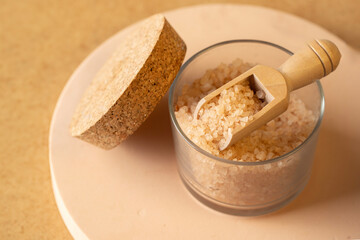 Cosmetic sea salt for relax bath in glass jar and wooden spoon on the podium beige background.