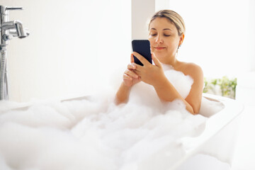 Young woman holds phone and takes bubble bath. Cute blonde is enjoying hot water and chatting on internet. Charming girl is looking at mobile screen and relaxed at home.