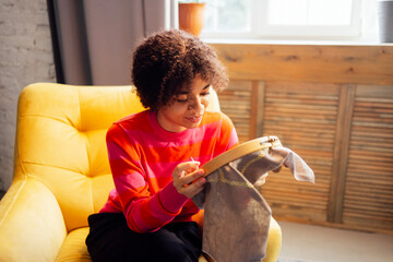A smiling young African woman in a red sweater is embroidering while sitting in an armchair