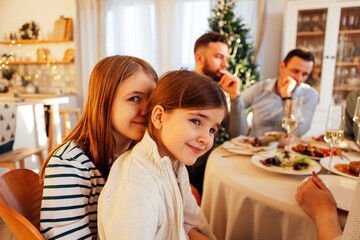 A cute teenage girl with her little sister at a festive table in the living room. A happy family celebrates Christmas together. Sweet smiling children have lunch with their parents.