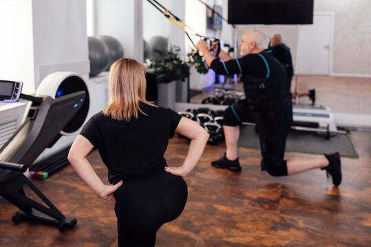 Elderly male athletic in electrical muscle stimulation suit uses trx system for his workout with personal trainer