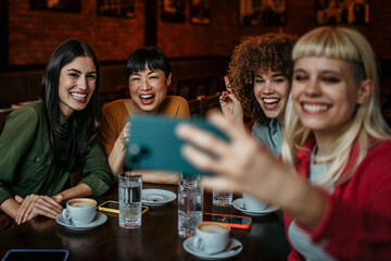 A young group of people sitting around a cafe table, taking a group selfie and drinking coffee.