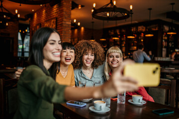 A young group of people sitting around a cafe table, taking a group selfie and drinking coffee.