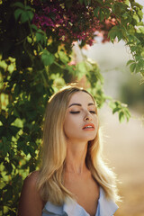 Fototapeta premium Beautiful blonde woman with closed eyes enjoying sunlight, surrounded by lush green leaves and purple flowers. Perfect for concepts of relaxation, beauty, and nature, capturing a serene moment.