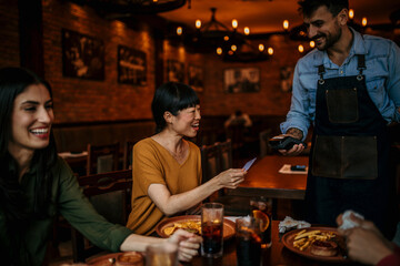 Mid aged Asian woman paying contactless using a credit card in the bar, with friends. A customer paying through credit card using contactless technology