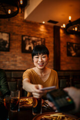 A group of friends is sitting in a coffee shop and a smiling woman is paying the bill with a credit card