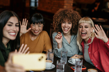 A multiracial group of young people sitting in a coffee shop, taking a group selfie and smiling.
