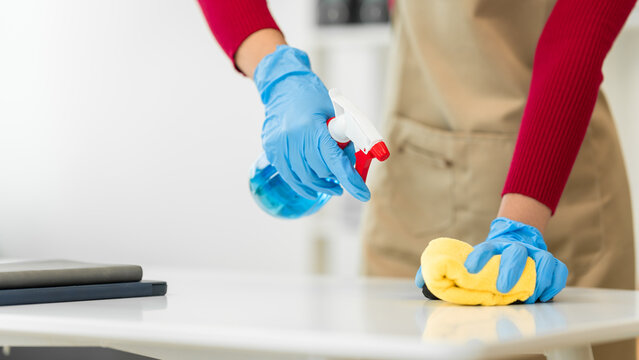 A young woman cleans her desk at home, using a disinfectant spray and a wipe. She ensures a hygienic and organized workspace, emphasizing cleanliness and personal health in her routine