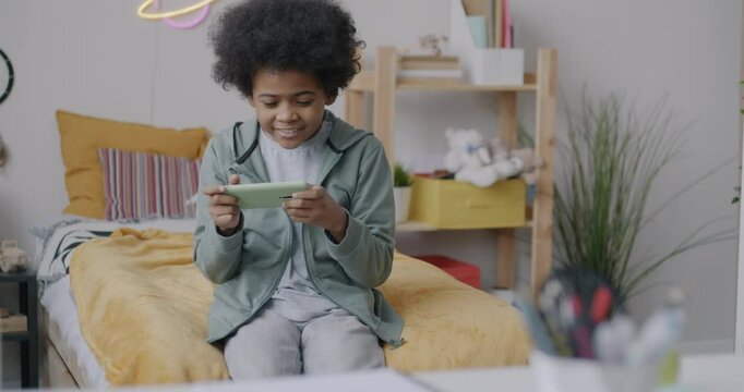 Joyful African American boy playing online video game with smartphone sitting on bed in bedroom. Gaming and childhood concept.