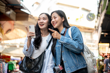 Two excited Asian female friends enjoy exploring a local market together, pointing at a food stall.