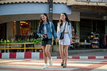 Two joyful young Asian women cross a crosswalk together while strolling through a city in Thailand.