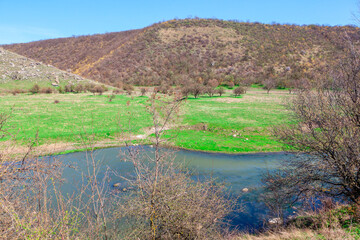 Scenic landscape featuring small river, surrounded by green grass and sparse trees. In the background there are hills and some rocky areas