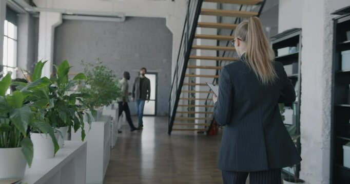 Back view dolly shot of businesswoman in suit walking in office reading document while people working around. Business and workplace concept.