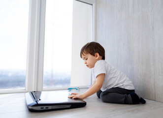 Young child playing with laptop near bright window. A young boy is sitting on the floor, engaged with a laptop beside a large window during daylight.