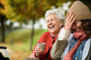 Portrait of a granddaughter and grandmother sitting on bench in autumn park, drinking coffee and...