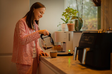 A young woman prepares coffee and tea in the kitchen, enjoying a peaceful moment. Perfect for themes of home life, kitchen activities, and relaxation