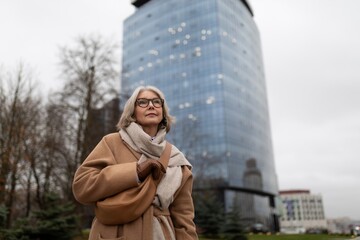 A woman in a beige coat walks thoughtfully past a modern glass building on a cloudy day in an urban setting