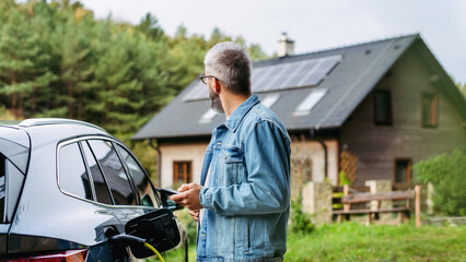 Man charging electric car in front of his house, plugging the charger into the charging port. House...