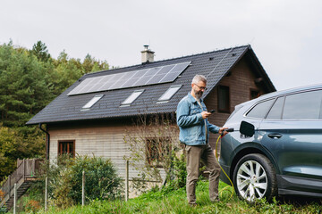 Man charging electric car in front of his house, plugging the charger into the charging port. House...