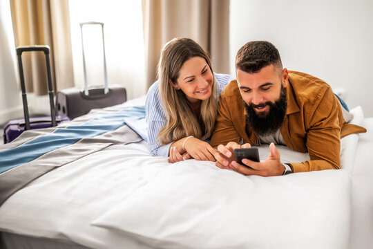 Young happy couple is on vacation. Tourists arrived in hotel room with their suitcases. They are lying on bed and using smartphone. - Powered by Adobe