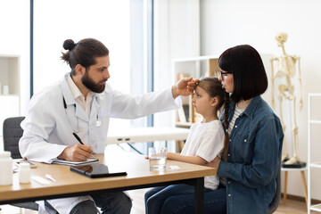 Caucasian male doctor measures temperature of young girl with concerned mother by her side in clinic. Doctor writes prescription while ensuring child's health and comfort.
