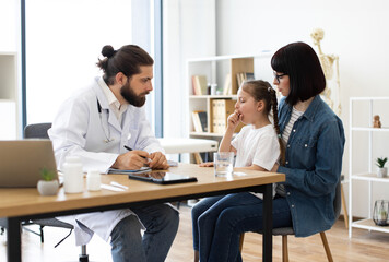 Caucasian male doctor examines young Caucasian girl coughing while mother listens in clinic. Doctor writes prescription as part of medical care.