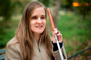 A visually impaired woman with a white cane sitting on a bench in a park on an autumn day