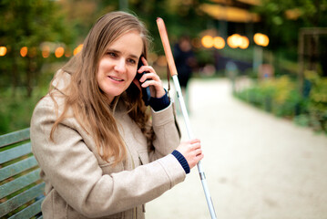 A visually impaired woman with a white cane sitting on a bench in a park on an autumn day and using...