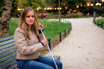 A visually impaired woman with a white cane sitting on a bench in a park on an autumn day