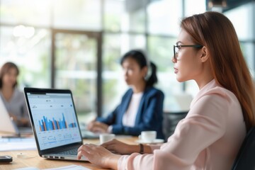 Obraz premium A focused female professional analyzing bar chart data on a laptop in a bright, modern office environment, with colleagues working in the background.
