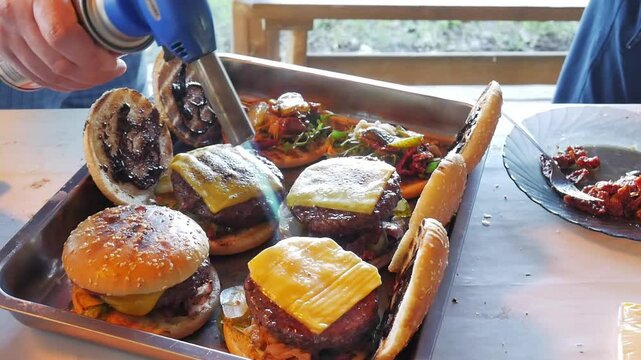 Making burgers and hamburgers outdoor.  Two friends preparing cheesebugres. One of them puts ham down and the other grills cheese slices with gas burner