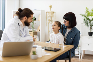 Caucasian male doctor interacts with young girl and mother in modern clinic setting. Female child smiles during consultation creating warm caring atmosphere. Doctor appears attentive and professional.