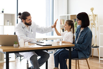 Obraz premium Caucasian male doctor gives high five to young girl sitting on Caucasian mother lap in clinic setting. Scene conveys support, trust, and care, highlighting positive doctor-patient relationship.
