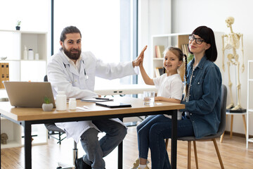 Caucasian male doctor gives high five to young girl sitting on mother's lap in clinic. Scene conveys trust and care in healthcare setting. Concept of family support and friendly physician interaction.