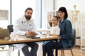 Male doctor interacting with young girl and mother in medical office. Image features Caucasian adult male doctor, Caucasian young girl with braids, and Caucasian adult female mother.