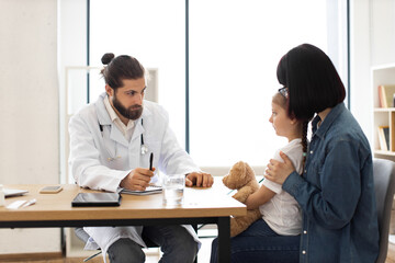 Caucasian male doctor examines young girl coughing in clinic while mother watches. Doctor writes down prescription, ensuring detailed care. Scene reflects healthcare and family wellbeing atmosphere.