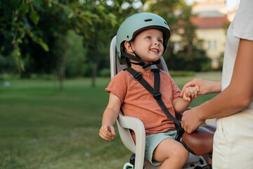 Portrait of an adorable boy wearing a helmet, sitting on rear bicycle child seat in a park looking with love at his mom. Space for text