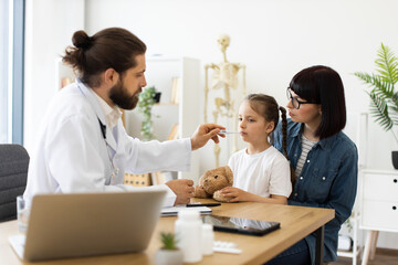 Caucasian male doctor measures temperature of little sick Caucasian girl sitting with mother....