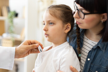 Caucasian doctor measures temperature of sick little girl using digital thermometer. Child appears calm while being comforted by Caucasian mother.