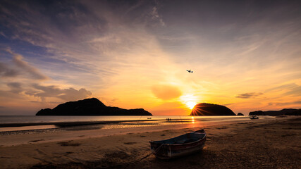 beautiful sunrise morning scenery, silhouette mountain and small tourist with flying passenger aircraft, the front scene is a fishing boat on the beach. wide angle view shot.