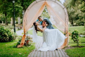 Bride and groom share a romantic dance pose under a decorative arch, surrounded by greenery in a...
