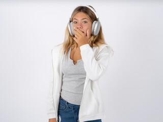 Beautiful teen girl wearing white jacket covers mouth and looks with wonder at camera, cannot believe unexpected rumors.