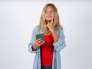 Beautiful teen girl wearing denim jacket thinks deeply about something, uses modern mobile phone, tries to made up good message, keeps index finger near lips.