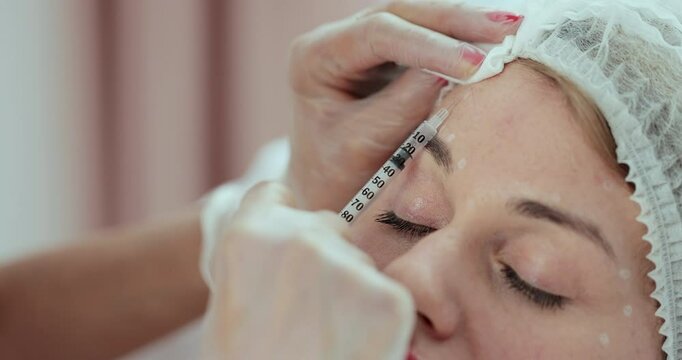 Cosmetologist makes botox injection in the forehead. Beautician in gloves holds a syringe ready to make beauty injection into female forehead.