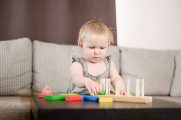 Baby plays logical wooden educational games sitting on couch. Wooden educational toys at home or in kindergarten. Concept of education, child development, leisure. Early childhood education, leisure