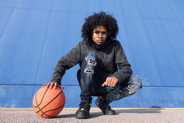 full body of serious african american teen male in informal outfit sitting with basketball ball...